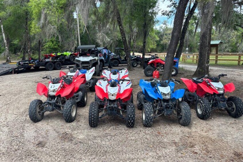 A row of quad bikes parked on a gravel surface with trees in the background. 4