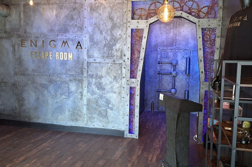 A metallic wall with the words 'ENIGMA ESCAPE ROOM' and a blue door with a light fixture above it. 4