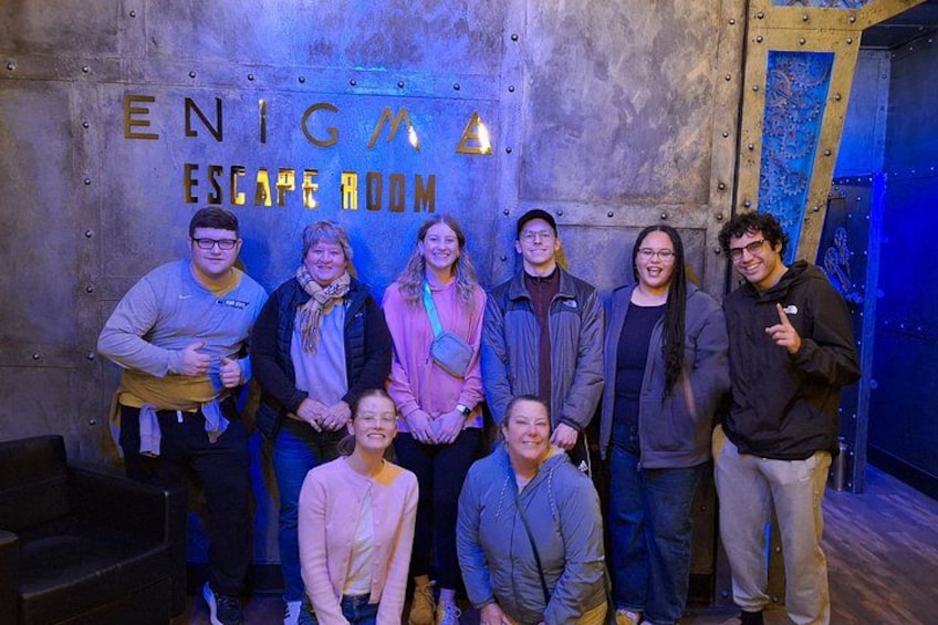 A group of people in front of a wall with the text 'ENIGMA ESCAPE ROOM'. 5