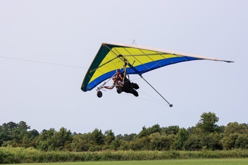 A person in a parachute harness gliding through the air over a grassy field with trees in the background. 2