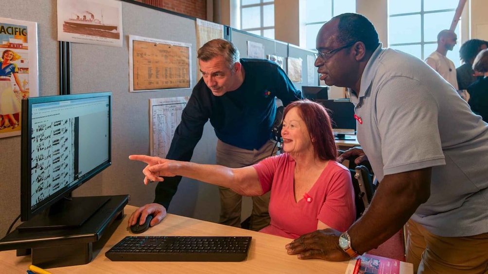 A person pointing at a computer screen, another person standing beside them, and a third person in a wheelchair. 2