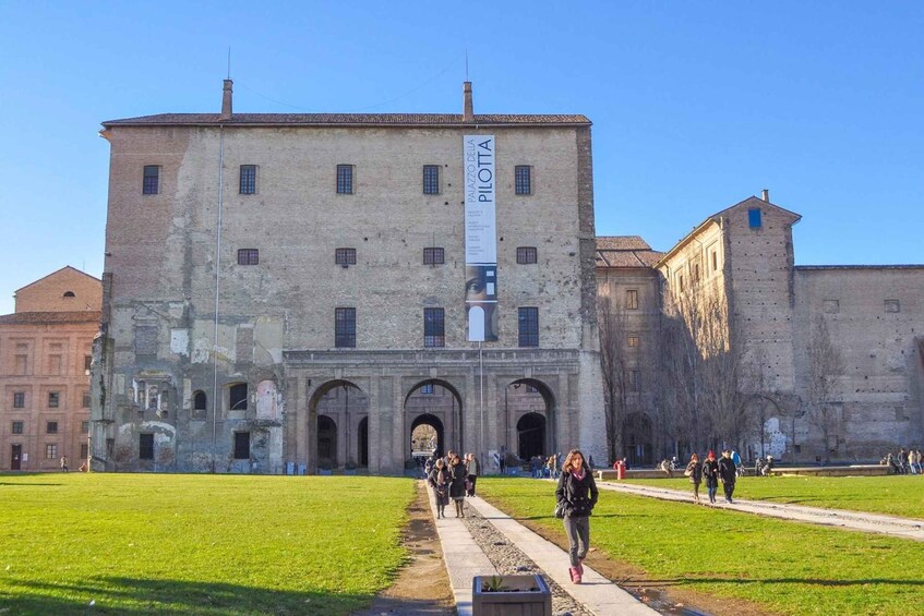 A historic building with arched entrances, a large banner, and a grassy courtyard. 3