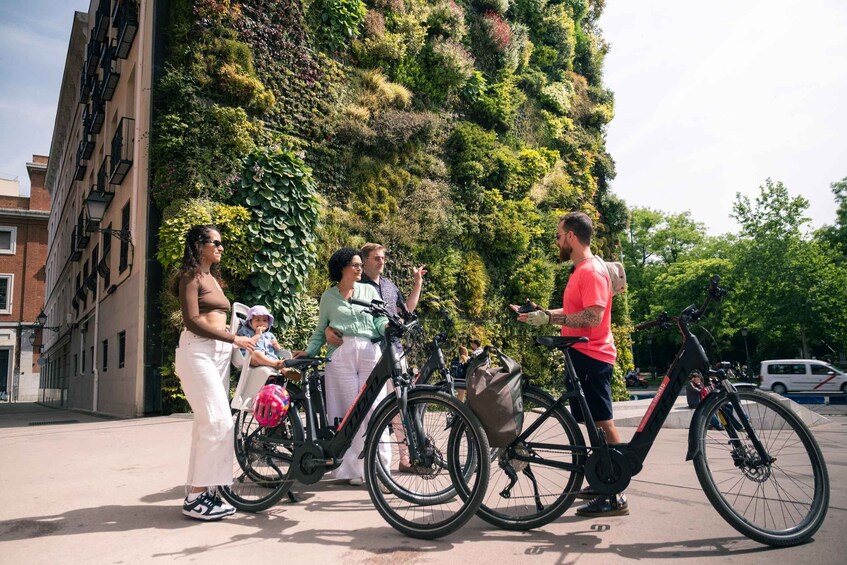 A group of people standing by bicycles in an urban setting with a green vertical garden wall in the background. 2