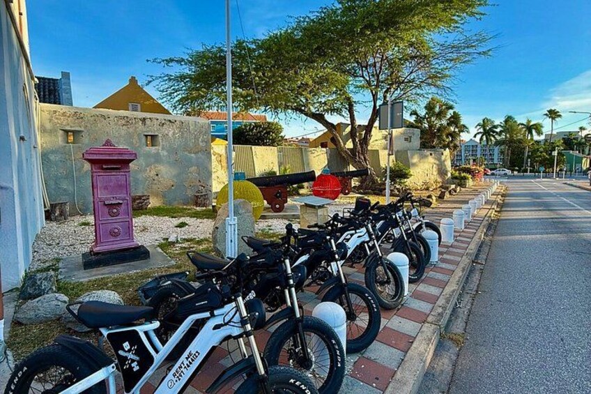 A row of bicycles parked along a sidewalk, a pink fire hydrant, and a building with a yellow roof in the background. 3