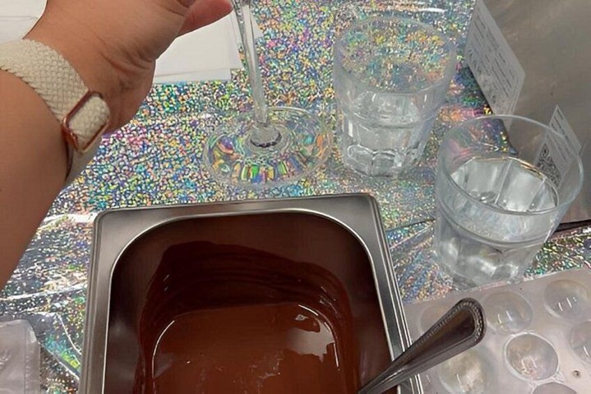A hand holding a glass, a metal container with a brown liquid, and two clear plastic cups on a glittery surface. 4