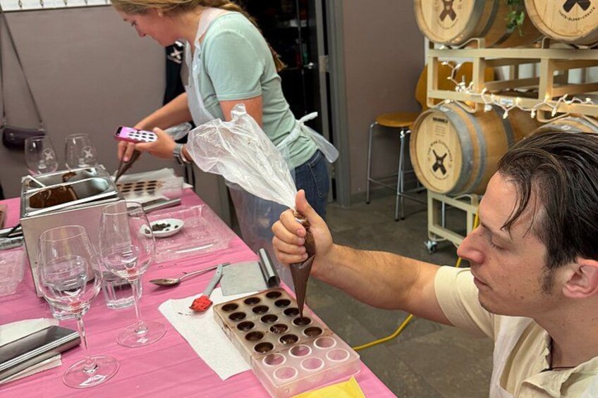 A person holding a plastic bag over a container with multiple holes, a pink table with wine glasses and a phone, and wooden barrels in the background. 5