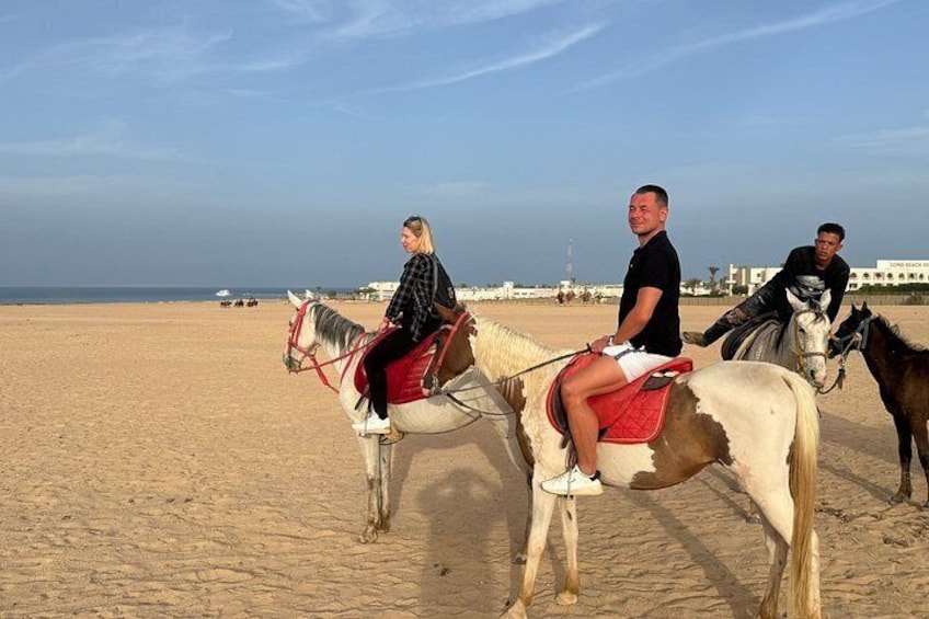Three people riding horses on a sandy beach with the sea and buildings in the background. 5