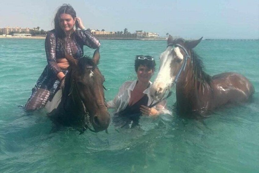 A person riding a horse in clear blue water with a beach and buildings in the background. 3