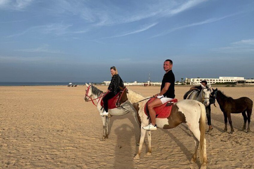 Two people riding horses on a sandy beach with a building in the background. 4