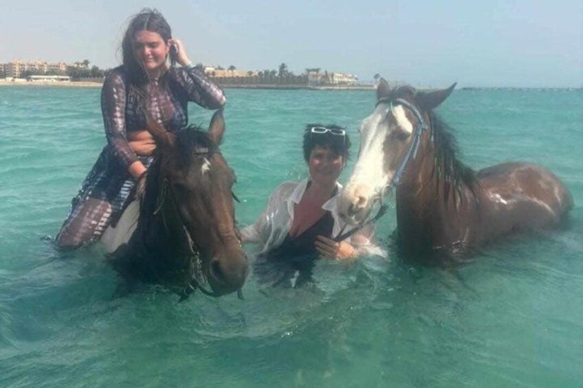 A person riding a horse in clear blue water with a cityscape in the background. 3