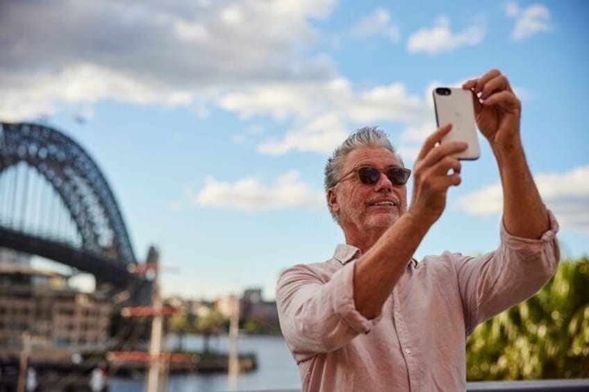 A person taking a photo with a smartphone, a large bridge, and a body of water in the background. 5