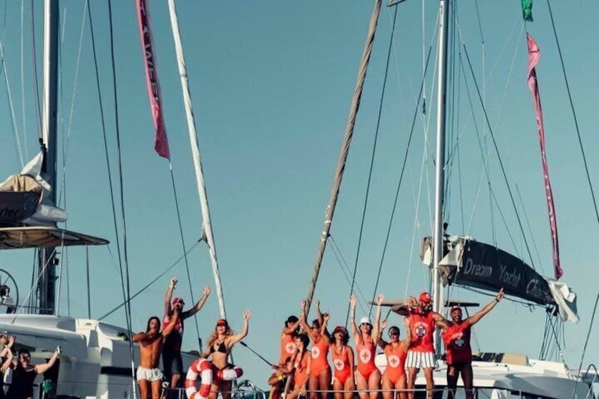 Un groupe de personnes se trouve sur un voilier, certaines d’entre elles portant des tenues assorties de couleur orange, et une banderole affiche l’inscription « Dream Yacht Club ». 5