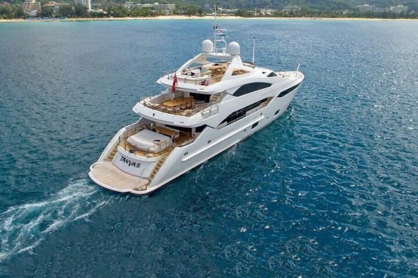 Un yacht blanc nommé « TANJAS » sur une étendue d’eau, avec une plage et des bâtiments en arrière-plan. 1