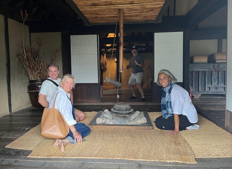 A group of people sitting on a woven mat in a traditional setting with a stone water feature and a bamboo ceiling. 3