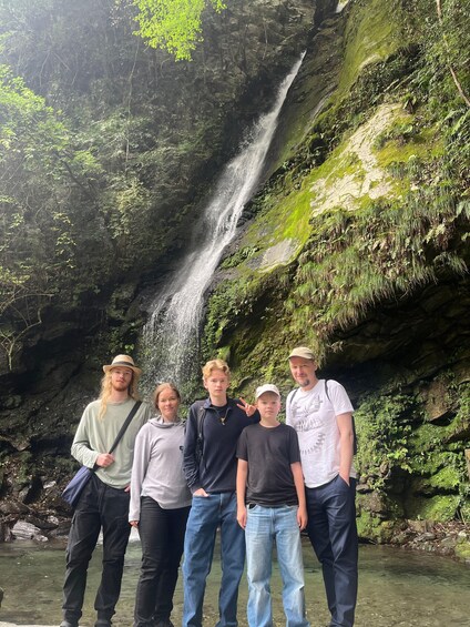 Un grupo de personas de pie frente a una cascada rodeada de exuberante vegetación y un risco rocoso. 4
