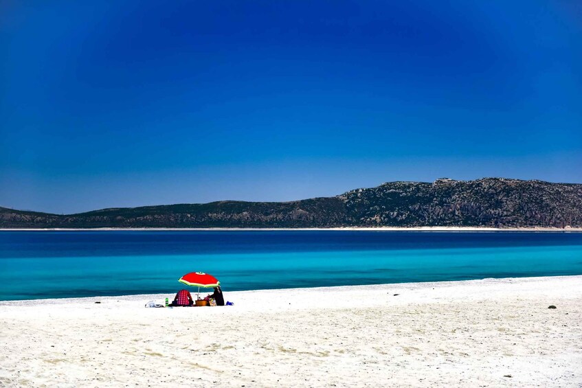 A beach with a person under a red and white umbrella, clear blue water, and a hilly landscape in the background. 5