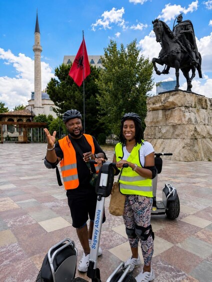 Two people on Segway scooters, a red flag, a statue of a horse, and a mosque in the background. 1