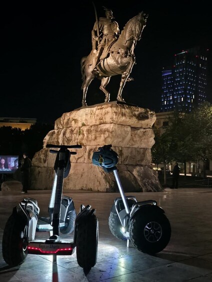 A nighttime scene with a statue of a horse and rider, two Segway scooters, and a modern building in the background. 5