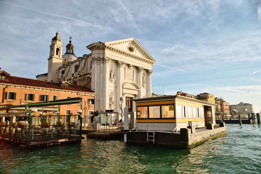 A canal with a boat docked, a historic building with a bell tower, and a restaurant with outdoor seating. 1