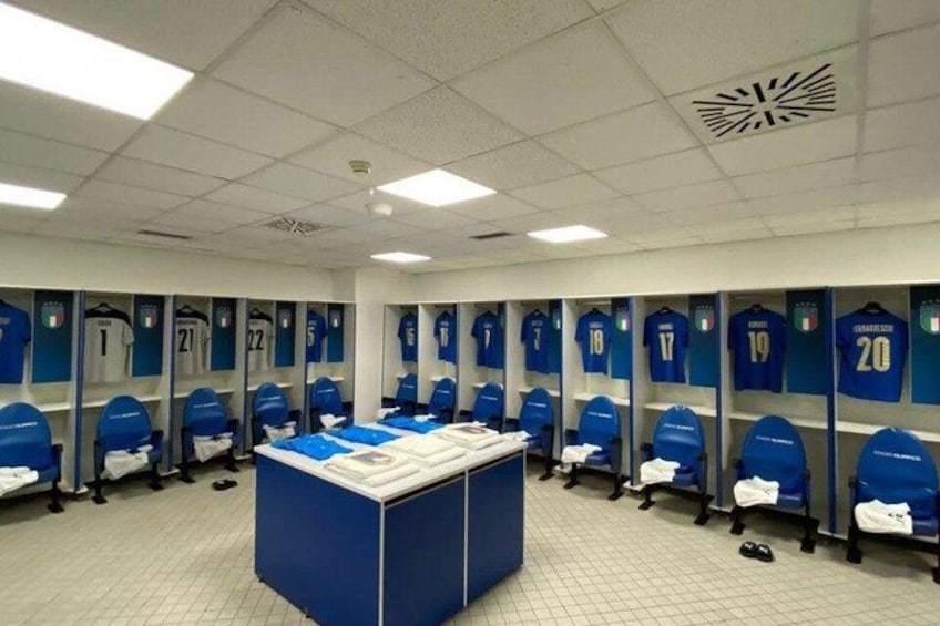A sports locker room with individual lockers numbered 1 to 20, a central table, and blue chairs. 4