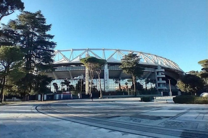A modern stadium with a large roof structure, surrounded by trees and a paved area. 2