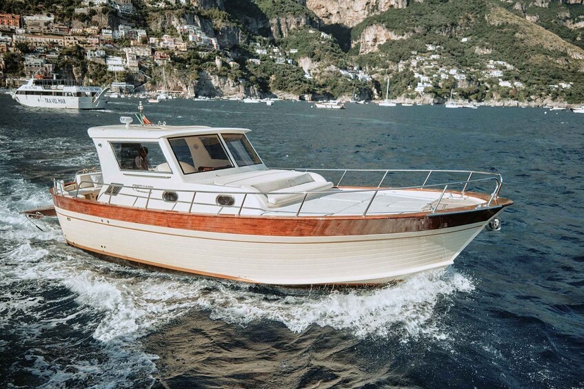 A white motorboat with a wooden deck and a cabin, moving through calm waters with a coastal town and cliffs in the background. 2