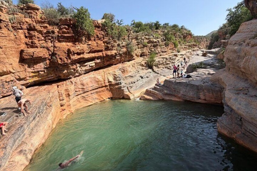 A natural pool surrounded by red rock formations with people swimming and standing on the edges. 5