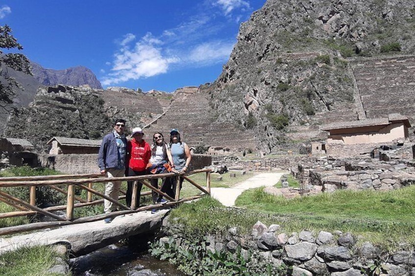 A group of people on a wooden bridge over a stream, with ancient ruins and a mountainous landscape in the background. 1