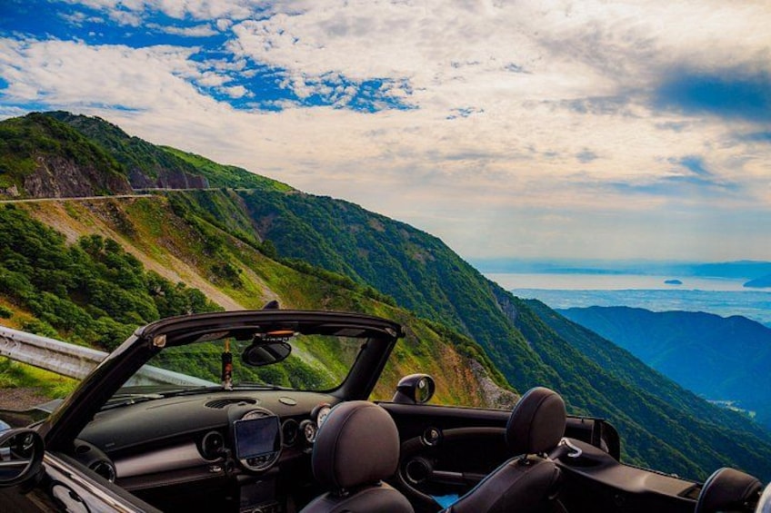 A convertible car driving on a mountainous road with lush greenery and a clear sky. 1