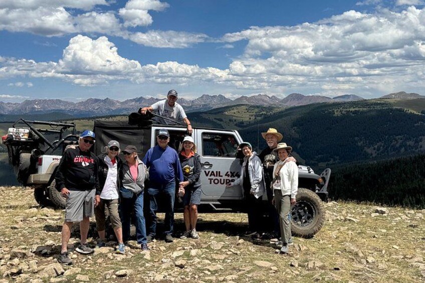 A group of people posing with a vehicle labeled 'RAIL 4X4 TOURS' on a rocky terrain with mountains in the background. 2