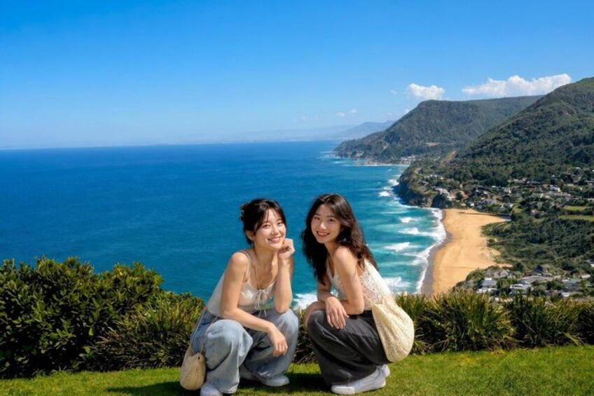 Two people sitting on a grassy hill overlooking a coastal landscape with a beach and hills in the background. 4