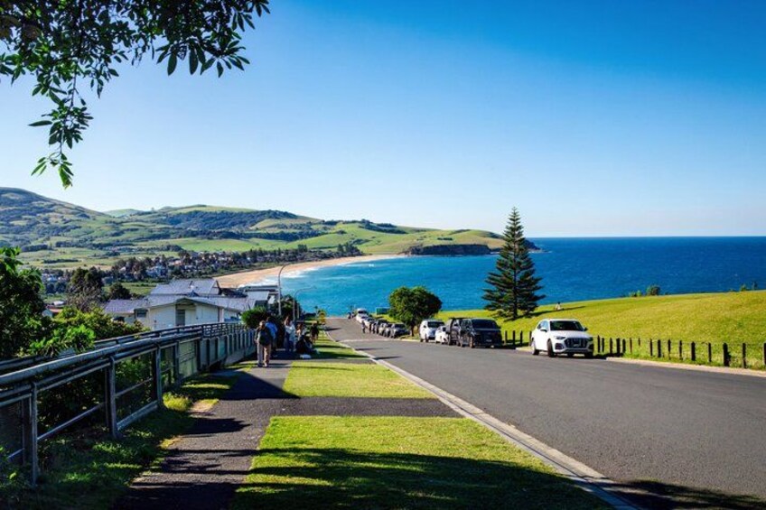 A coastal road with a grassy verge, a metal railing, and a clear view of the sea and distant hills. 2