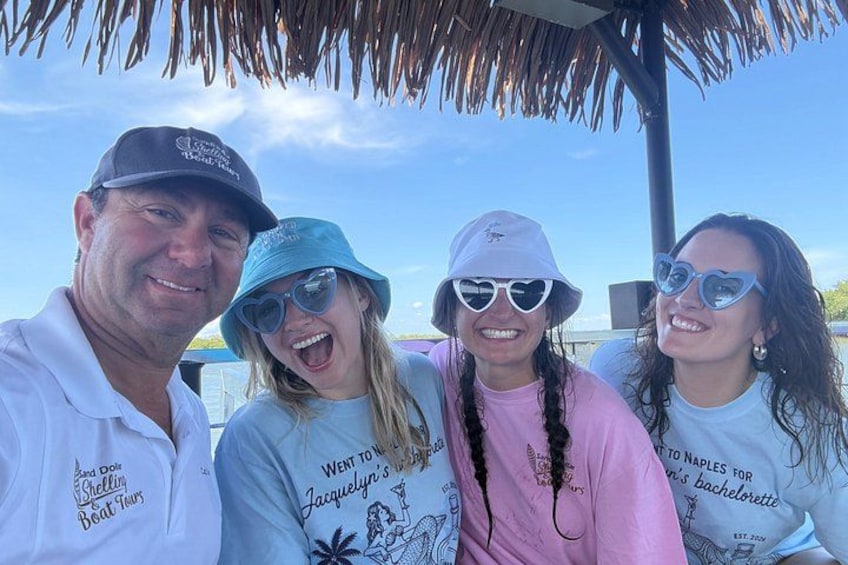 Four people wearing t-shirts with text, standing under a thatched roof structure. 1