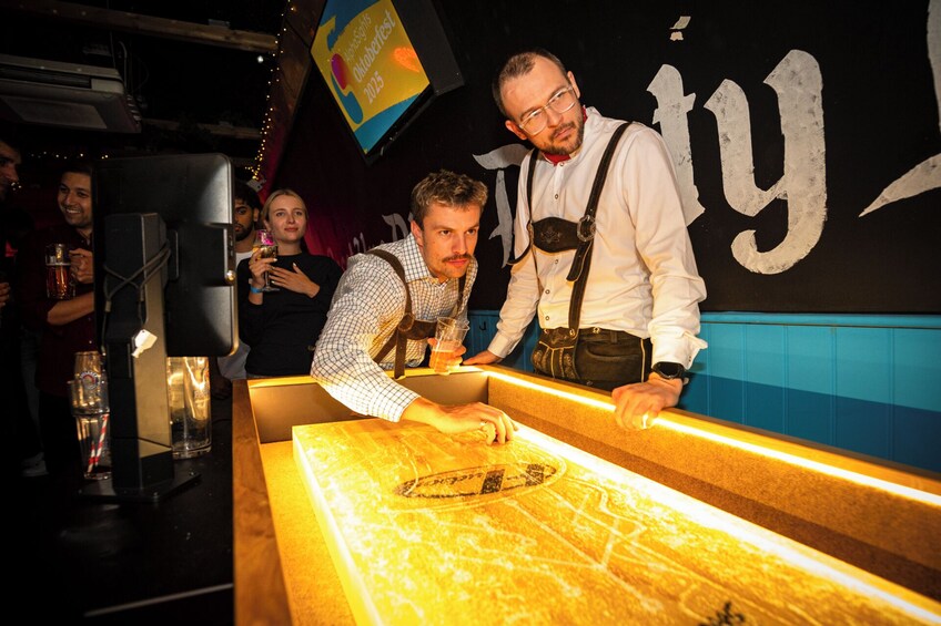 Two people at a bar counter with a sign reading 'Octoberfest 2025' in the background. 3