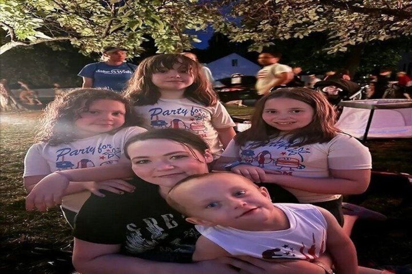 A group of people outdoors, some wearing t-shirts with text, under a tree with sunlight filtering through. 2
