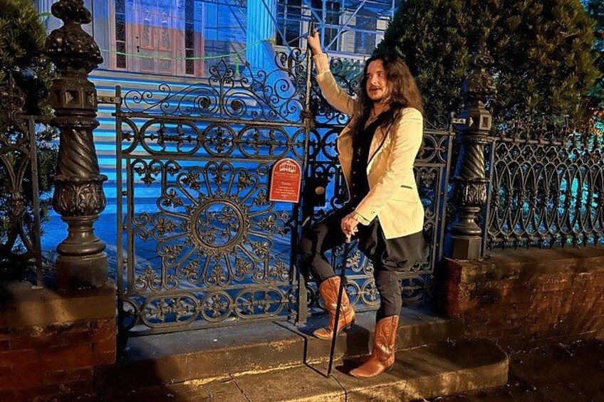 A person standing on steps next to a wrought iron gate with a sun emblem, wearing a beige jacket and boots. 2