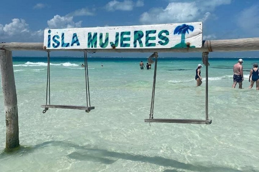 A beach with a sign reading 'Isla Mujeres' and clear turquoise water. 2