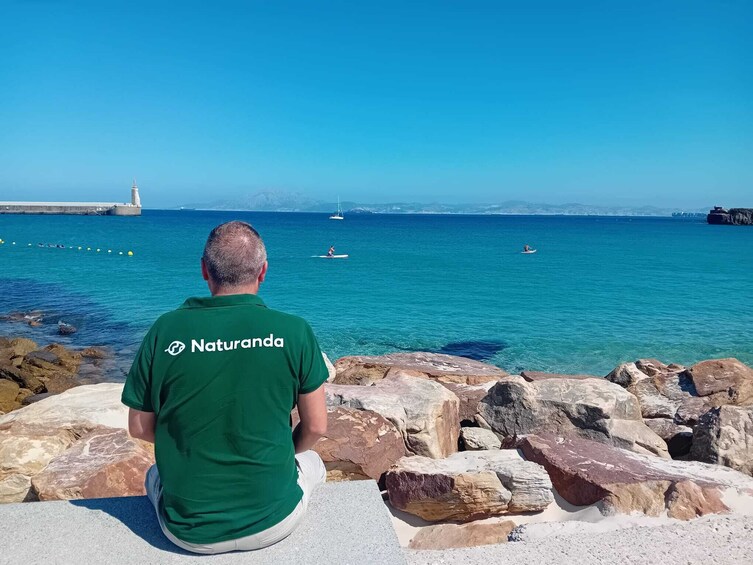Un homme vêtu d’une chemise verte, avec l’inscription « Naturanda » dans le dos, est assis sur des rochers au bord de la mer, avec des bateaux visibles au loin. 1