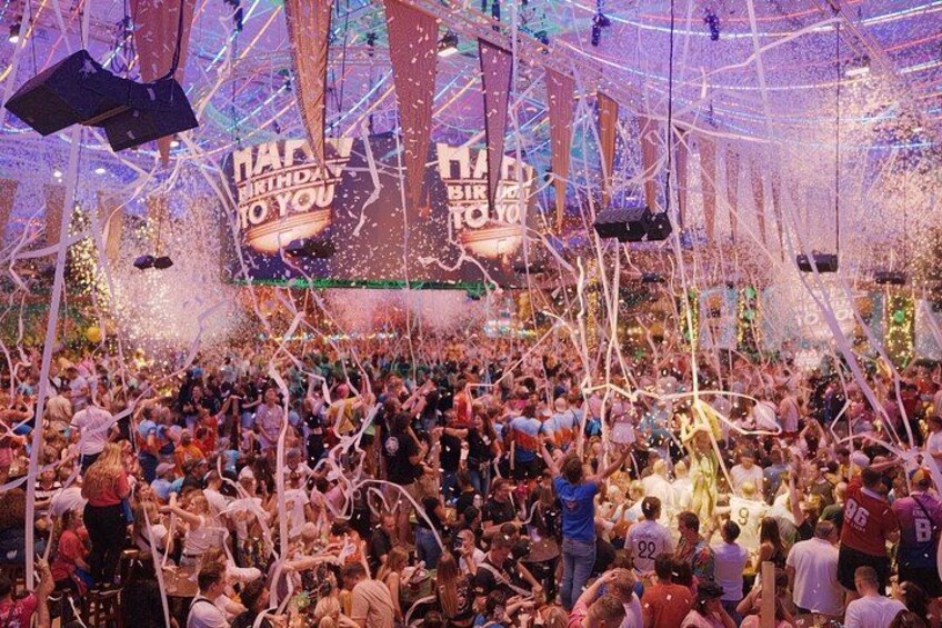 A crowded indoor event with confetti falling and a stage with a 'Happy Birthday' banner. 1