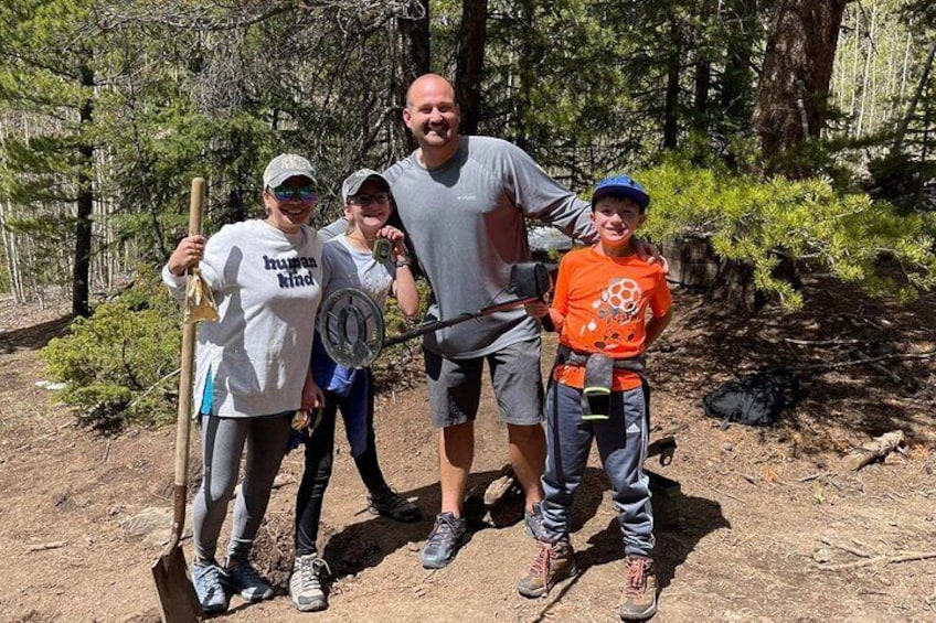 Four people in a forested area, one holding a shovel, another with a wheelbarrow, and two with backpacks. 1
