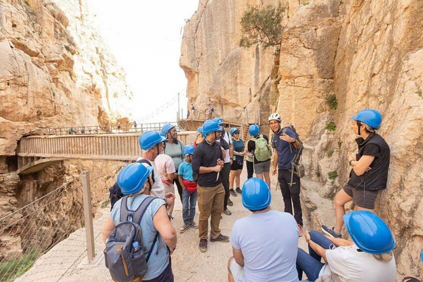 A group of people wearing helmets and backpacks, standing on a rocky path with a bridge in the background. 3