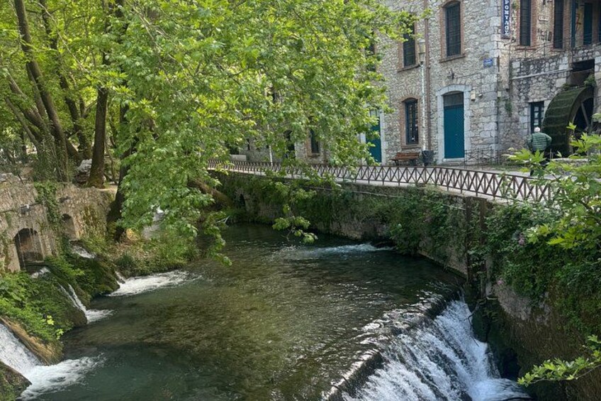 A stone bridge over a river with a waterfall, surrounded by lush greenery and historic buildings. 5