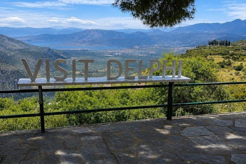 A stone bench with a sign reading 'VISIT DELPHI' overlooking a mountainous landscape. 1