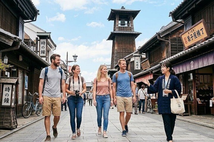 A group of people walking on a cobblestone street with traditional wooden buildings and a clock tower in the background. 1