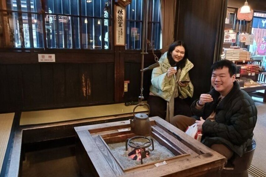 Two people sitting at a wooden table with a teapot and cups, in a room with traditional decor and a sign in Japanese. 2