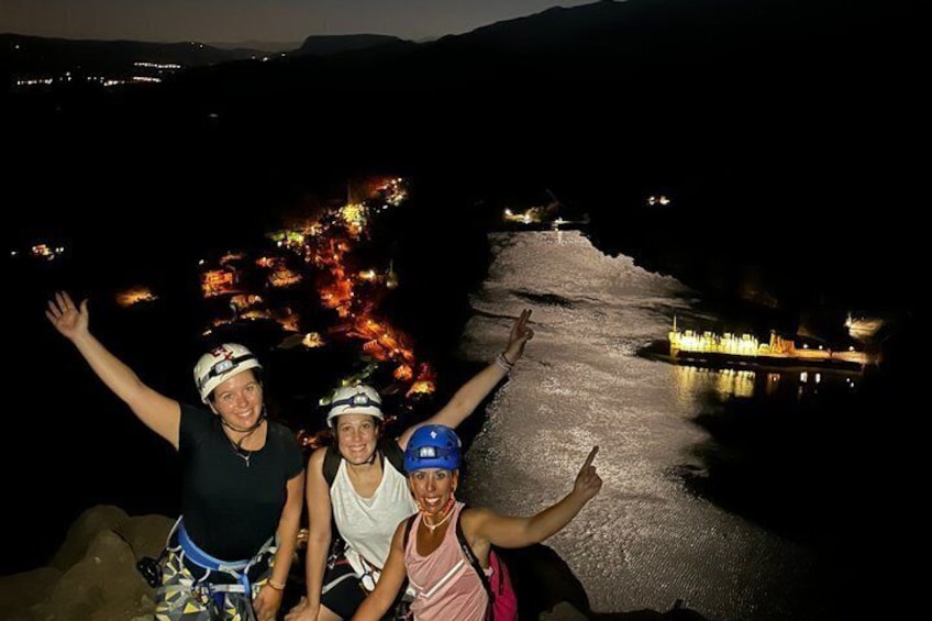 Three people with arms raised, overlooking a river with lights reflecting on the water, and a cityscape in the background. 3