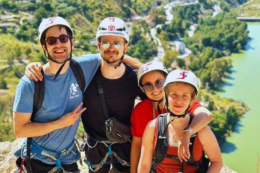 Four people wearing helmets and harnesses, standing on a cliff with a river and greenery in the background. 4