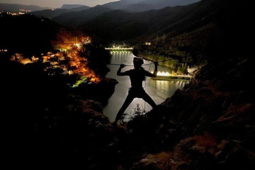 A person rappelling down a cliff with a river and illuminated buildings in the background. 4