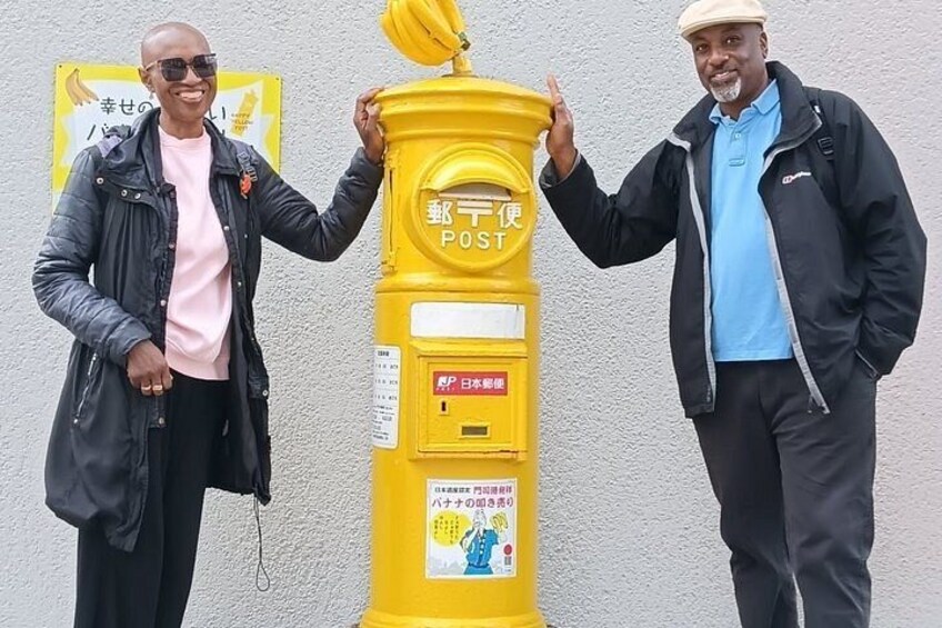Two men standing next to a yellow post box with Japanese text. 4