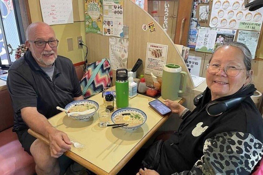 Two people sitting at a table with bowls of food, a green thermos, and a smartphone. 5